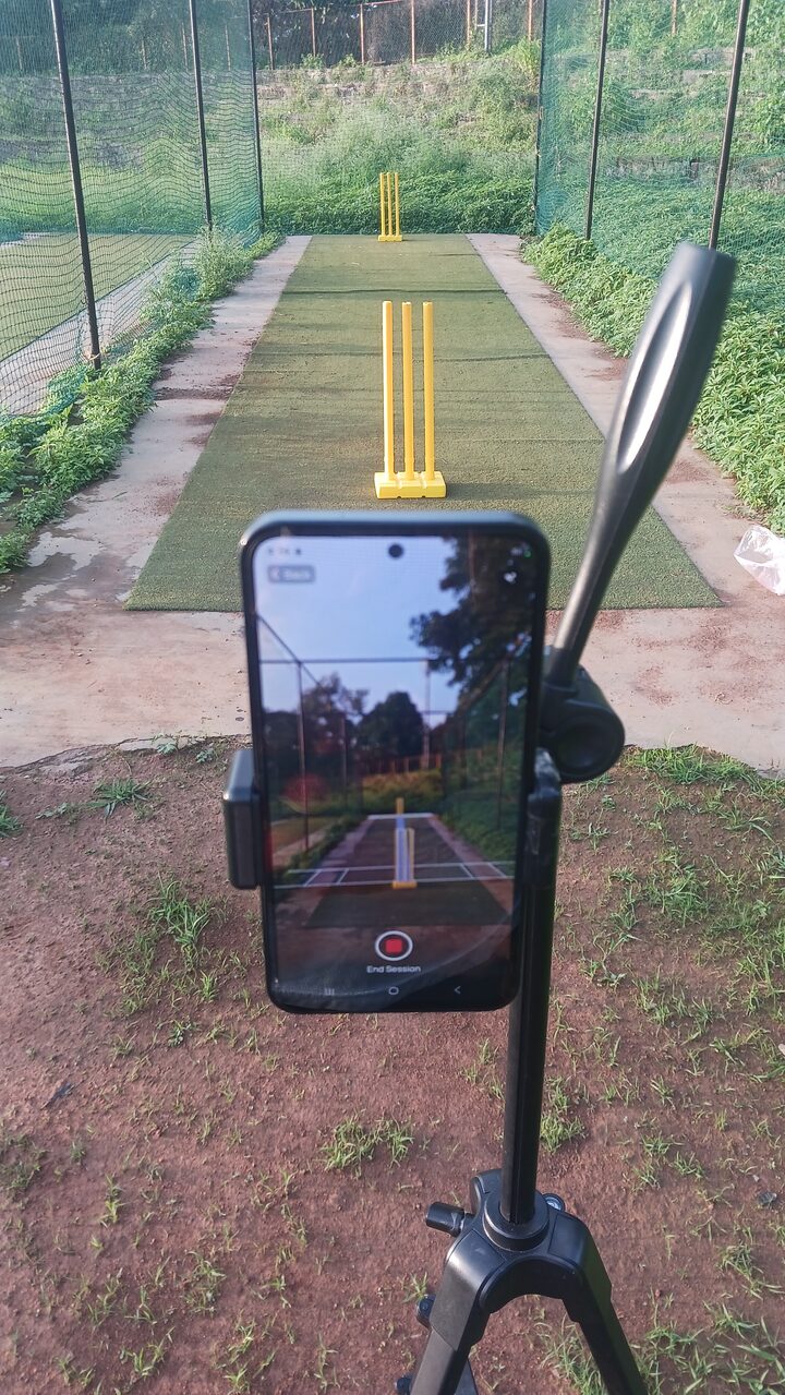 A phone mounted on a tripod behind the stumps in a cricket net, its screen showing Matcha recording the session down the pitch.