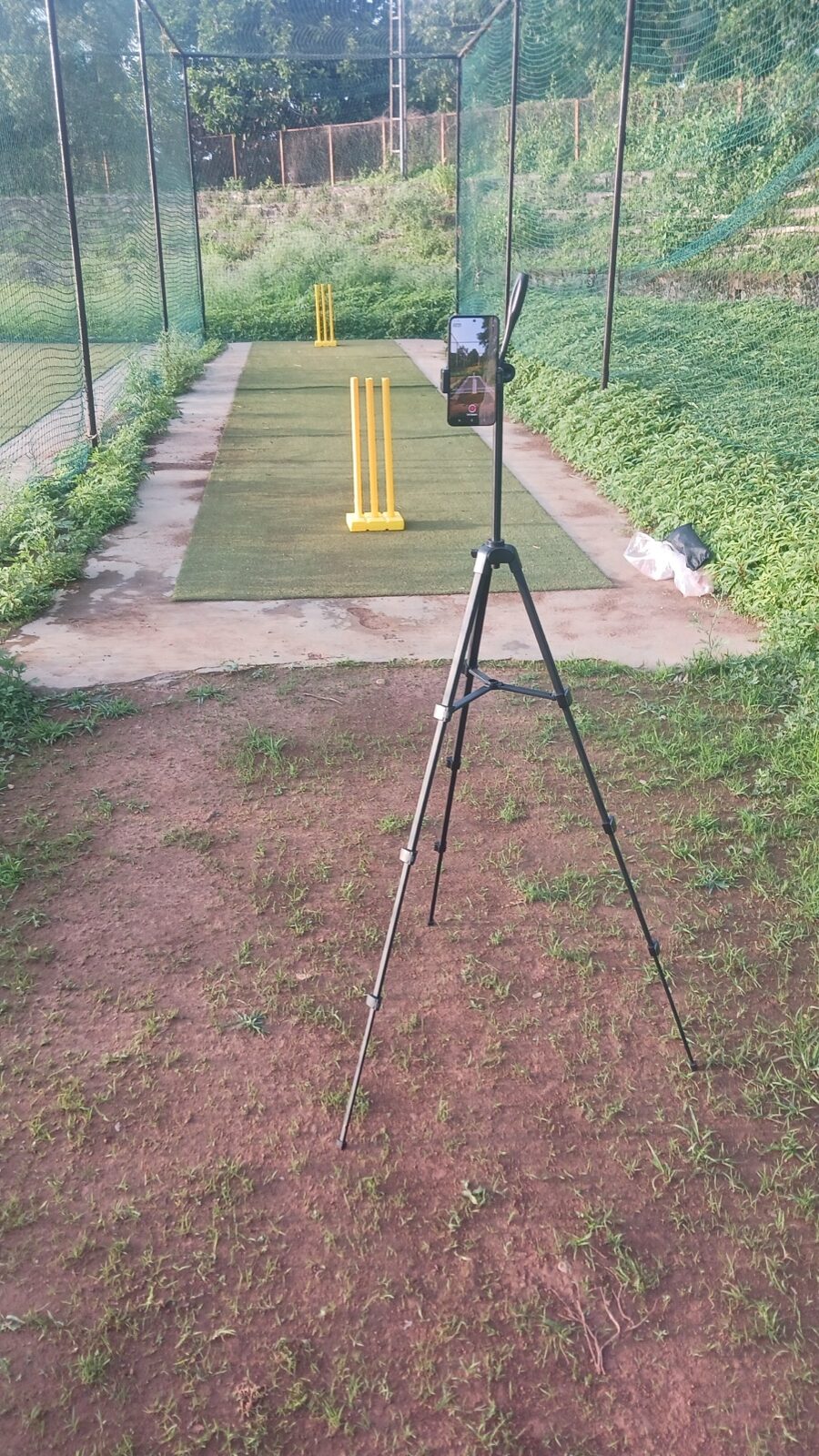 Phone mounted on a tripod behind the non-striker stumps in a cricket training net.